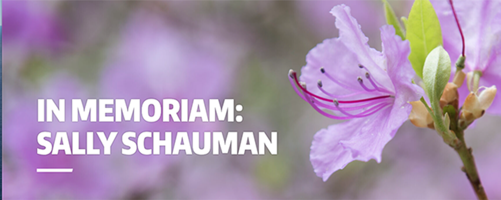 White text reading "In Memorium: Sally Schauman" on a blurred background with a closeup of a pink flower